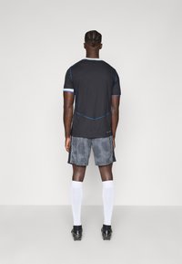 Kurzarm schwarzes Sportshirt mit blauen Akzentnähten und gemusterten grauen Shorts, kombiniert mit weißen Socken und schwarzen Fußballschuhen.