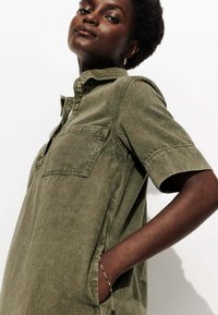 Femme confiante avec des cheveux bouclés courts portant une robe chemise verte olive à boutons avec une poche poitrine et un bracelet, les mains dans les poches.