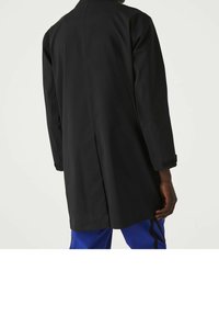Černý dlouhý kabát s rovný střih, s rozparkem na zadní straně, dlouhými rukávy a strukturovanou tkaninou. Nositý přes modré kalhoty s bočními akcenty.