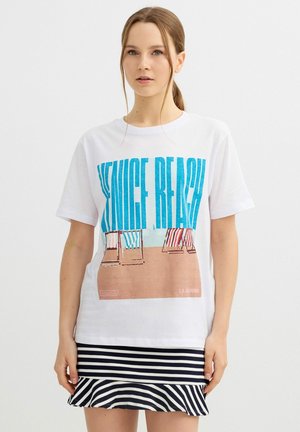 Joven mujer de cabello claro lleva una camiseta blanca con el texto "Venice Beach" y un gráfico de una silla de playa, combinada con una falda de rayas en blanco y negro.