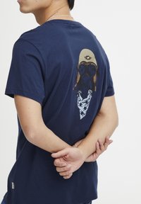 Temno modra bombažna majica s kratkimi rokavi, ki na hrbtni strani prikazuje grafični dizajn lika z očali in bandano. Reden kroj.