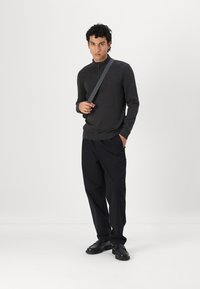 Pulover gri închis cu fermoar la gât, asortat cu pantaloni negri largi și pantofi negri. Modelul poartă o geantă gri subțire cu bandulieră. Fundal simplu.