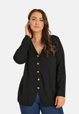 Kvinde med langt brunt hår iført sort knappet cardigan og blå jeans, poserer med den ene hånd nær øret.