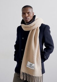 Beige uld tørklæde med frynser i enderne, draperet om halsen, båret over en mørk marineblå frakke. Har et hvidt mærke med kursiv tekst.