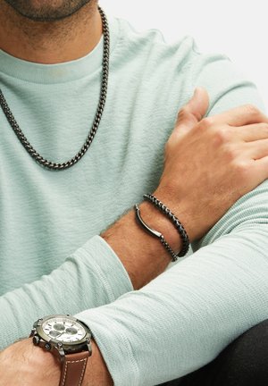 Silver chain necklace and black bracelet on a wrist. Light blue textured long-sleeve shirt and a black watch with a brown strap.