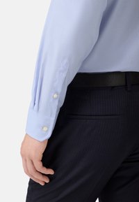 Svetlo modrá pruhovaná košeľa s dvoma gombíkmi na manžetách, nosená s tmavými pruhovanými nohavicami a čiernym pásom. Tkanina sa zdá byť hladká.