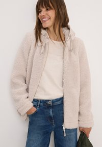 Femme souriante avec une main dans la poche, portant une veste zippée en sherpa beige sur une chemise crème et un jean bleu, tenant un objet matelassé vert.