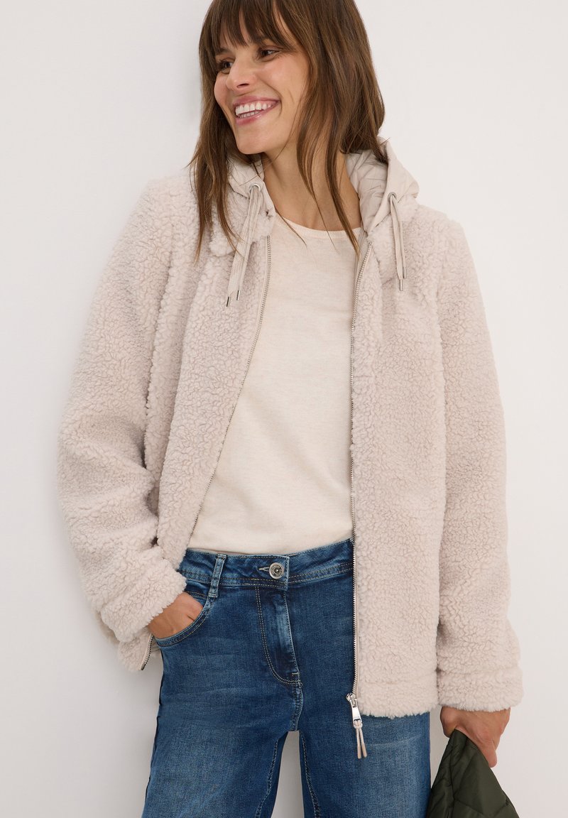 Femme souriante avec une main dans la poche, portant une veste zippée en sherpa beige sur une chemise crème et un jean bleu, tenant un objet matelassé vert.