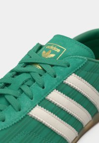 Πράσινο σουέντ αθλητικό παπούτσι με τρεις λευκές ρίγες, υφή στην επάνω πλευρά, στρογγυλά κορδόνια και χρυσό λογότυπο στη γλώσσα.