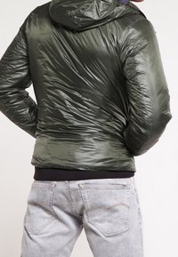 Olivengrøn pufferjakke med hætte, der har en blank, struktureret overflade og elastiske manchetter. Bæret over grå jeans med diskrete syninger.
