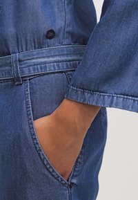 Džínový overal v modré barvě, s páskem v pase, dlouhými rukávy a boční kapsou. Černý detail knoflíku poblíž pasu.