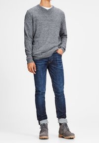 Grauer V-Ausschnitt-Pullover, blaue Jeans und graue Stiefel mit hellen Sohlen. Der Pullover hat ein strukturiertes Strickmuster; die Jeans sind eng geschnitten mit umgeschlagenen Säumen.