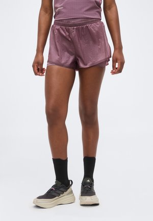 Person trägt mauvefarbene Sportshorts und Top, schwarze Socken sowie beige und schwarze Laufschuhe und steht vor einem weißen Hintergrund.