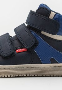Zapato alto para niño pequeño en azul y negro con correas de velcro, suela de goma, acentos de tela texturizada y un logo rojo de Kickers.