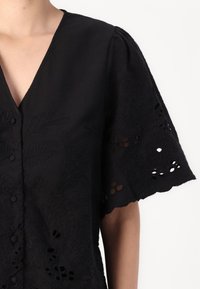Black blouse with short, puffed sleeves featuring embroidered leaf patterns and eyelet details. V-neckline with button closure.
