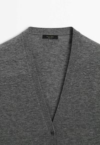 Cardigan gris tricoté avec col en V et un seul bouton visible, montrant la texture du tissu et une étiquette noire à l'intérieur du col.