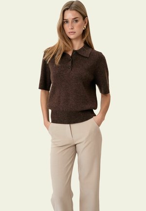 Femme aux cheveux châtain clair lisses, portant un polo tricoté à manches courtes marron foncé et un pantalon beige, debout les mains dans les poches.