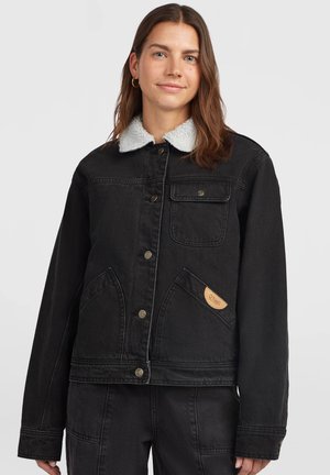 Femme portant une veste en jean noire avec un col sherpa blanc, des fermetures à boutons et des poches avant, debout devant un fond clair uni.