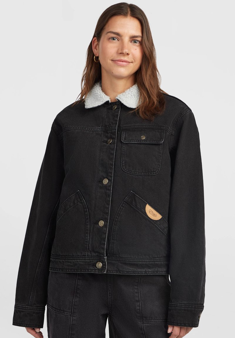 Жена, носеща черно дънково яке с бял яка от шерпа, закопчаване с копчета и предни джобове, стояща пред обикновен светъл фон.