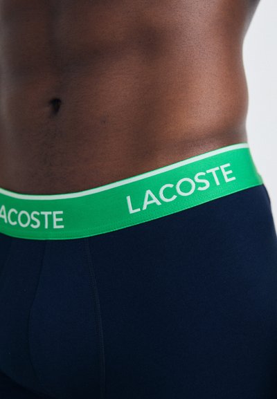 Тъмносини боксерки с зелен колан и бяло лого "LACOSTE". Тъканта изглежда мека и еластична.