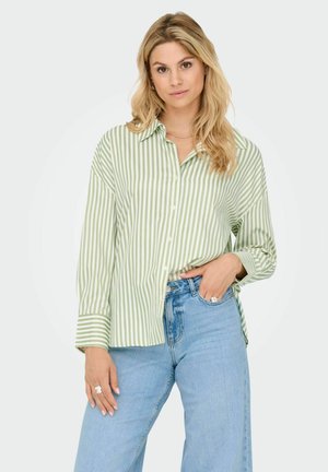 Blond kvinna iförd en grön och vit vertikalt randig skjorta med knappar och ljusblå högmidjade jeans, stående med en hand i fickan.