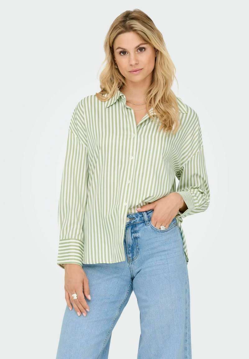 Femme blonde portant une chemise à rayures verticales vertes et blanches et un jean taille haute bleu clair, se tenant avec une main dans la poche.