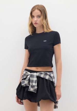 Zwart gekropte t-shirt met een wit 'oops' logo, gecombineerd met een zwarte uitlopende rok en een geruite flanellen shirt dat om de taille is geknoopt.