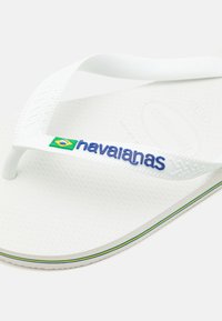 Vit flip-flop i gummi med strukturerad sula. Har vita remmar med "havaianas"-logotyp i blått och en grön och gul accent.