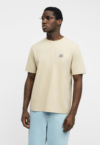 Άνδρας που φοράει μπεζ κοντομάνικο μπλουζάκι με μικρό λογότυπο λύκου στο στήθος και ανοιχτό μπλε τζιν, κοιτάζοντας προς το πλάι μπροστά σε απλό φόντο.