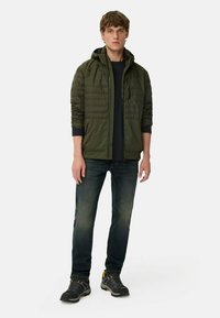 Olivgrüne gepolsterte Jacke mit Kapuze, Reißverschluss und elastischen Bündchen, kombiniert mit dunkelblauen Jeans und grauen Sneakers mit gelben Akzenten.