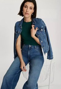 Jeansowa kurtka w średnim niebieskim kolorze z srebrnymi guzikami, noszona na ciemnozielonym topie; zestawiona z wysokim stanem, szerokimi niebieskimi dżinsami.