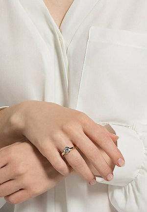 Goldring mit einem runden, klaren Stein, glatter Band, getragen an einer Hand, die über einer weißen Bluse mit einer geknöpften Brusttasche ruht.