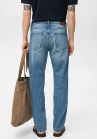 Lichtblauwe denim jeans met een rechte pasvorm, vijf zakken en een leren label aan de achterkant. Een bruine canvas tas wordt in de linkerhand vastgehouden.