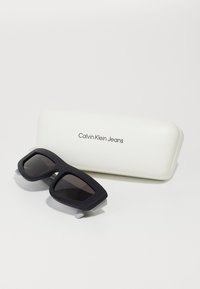 Calvin Klein Jeans juodi akiniai nuo saulės su stačiakampiais rėmeliais, tamsiais lęšiais ir matinėmis tekstūromis, padėti šalia balto dėklo su reljefiniu logotipu.
