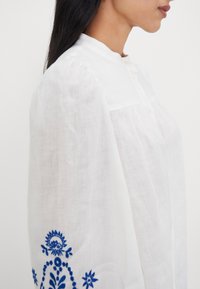 Femme portant un chemisier blanc avec une broderie florale bleue sur la manche, debout de profil devant un fond uni.