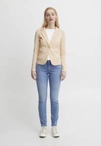 Světlobéžový upravený blazer s jedním knoflíkem, dvěma předními kapsami, kombinovaný s bílým tričkem, modrými džínami a bílými teniskami.