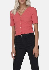 Cardigan tricot à manches courtes couleur corail avec un col festonné, une fermeture à boutons et un ourlet festonné décoratif. Porté avec un jean noir taille haute.