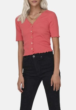 Cardigan tricot à manches courtes couleur corail avec un col festonné, une fermeture à boutons et un ourlet festonné décoratif. Porté avec un jean noir taille haute.