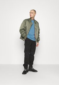 Olivgrüne Bomberjacke mit Rippbündchen, darunter ein blaues T-Shirt, schwarze Cargo-Hosen und schwarze Sportschuhe mit grauen Schnürsenkeln.