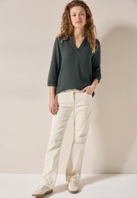 Blusa lunga a maniche lunghe di un verde scuro texture, con scollatura a V, abbinata a jeans dritti color crema e sneakers chiare. Design semplice.