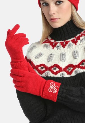 Gants en tricot rouges avec des poignets côtelés, présentant un logo décoratif au poignet. Le modèle porte un pull noir avec un col épais et à motifs.