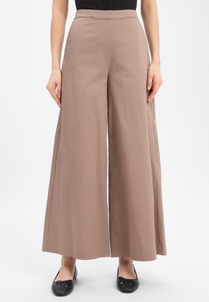Beige weite Hosen, getragen mit schwarzen Ballerinas, bei einer Person, die vor einem weißen Hintergrund steht.