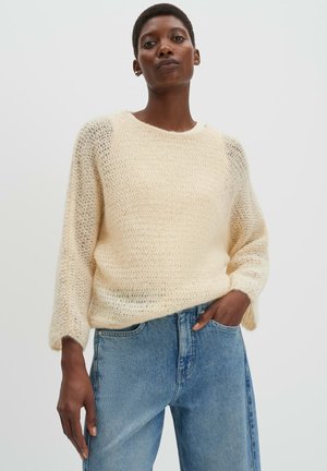 Frau trägt einen cremefarbenen, locker gestrickten Pullover und hellblaue, hoch taillierte Jeans, steht vor einem schlichten hellen Hintergrund.