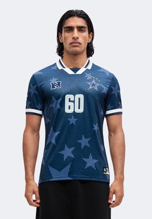 Jonge man met donker haar draagt een blauw sportshirt met witte sterren en nummer 60, staand tegen een effen lichte achtergrond.