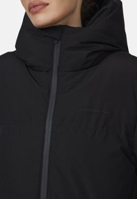 Giacca a vento nera con collo alto, dotata di chiusura centrale con zip e logo in rilievo. Il materiale appare liscio con sezioni imbottite per l'isolamento.