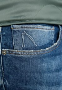 Jeans in denim con un lavaggio blu, con una tasca frontale decorata con un logo cucito e hardware in colore rame. Bordo che mostra una leggera usura.
