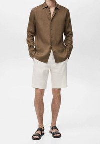 Camicia di lino marrone con chiusura a bottoni, abbinata a pantaloni corti di lino bianchi e sandali neri. Silhouette semplice e rilassata con texture naturali.