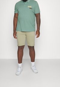 Polo verde con logo, abbinato a pantaloni corti beige arrotolati. Sneakers bianche e calzini completano il look. Tessuto morbido e design casual.