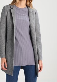 Abrigo de mezcla de lana gris con un diseño estructurado y solapas de pico, llevado sobre una blusa de color lavanda con canalé, combinado con vaqueros azul oscuro.