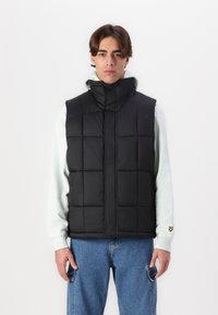 Black puffer vest with a high collar, featuring a quilted design. Worn over a light gray hoodie and paired with blue jeans.